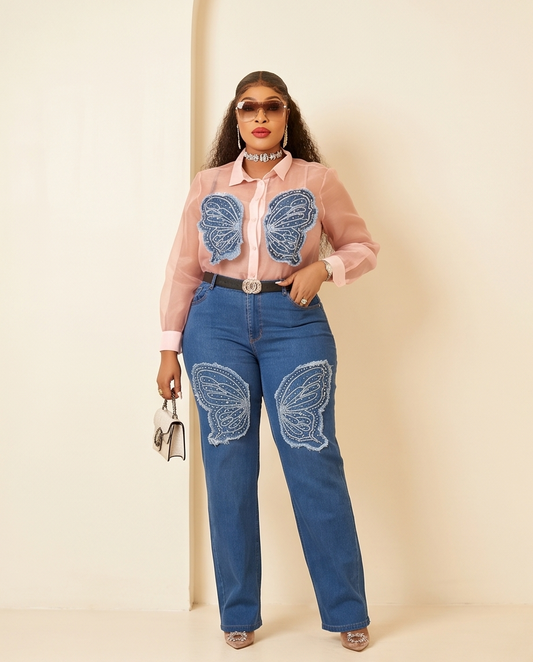A woman models a pink sheer long-sleeve button-up shirt with blue butterfly appliques on the chest, paired with blue jeans featuring matching butterfly patches on the thighs, styled with a black belt, sunglasses, and a small handbag against a cream background.