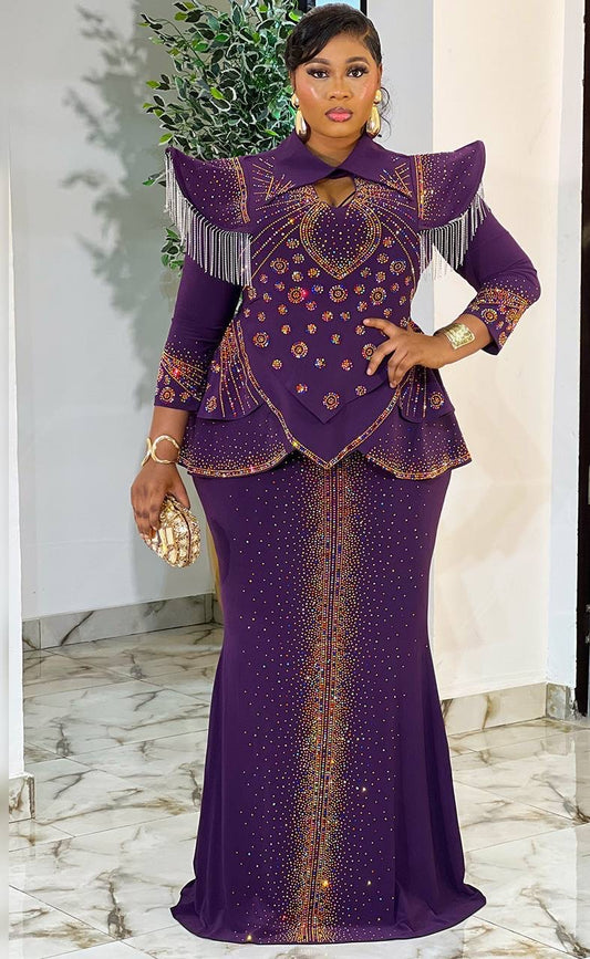 A woman models a purple traditional African dress with gold embroidery, fringe details, and a flared peplum top, standing in an indoor setting with a marble floor.
