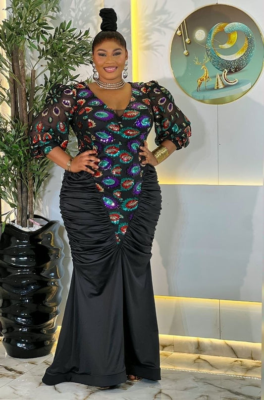 A woman modeling a floor-length black mermaid gown with a heavily embellished multicolor bodice featuring red, green, blue, and purple fish-scale patterns, posed in an indoor setting with a plant and decorative wall art.