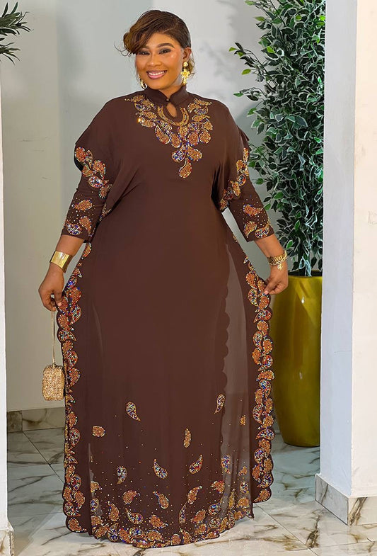 A woman models a brown long-sleeve maxi dress with intricate multicolor floral and paisley embroidery, standing in an indoor setting with a potted plant in the background.