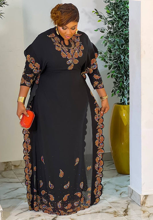 A woman wearing a black maxi dress with orange and brown paisley and floral embroidery, holding a small orange clutch in an indoor setting with a potted plant.