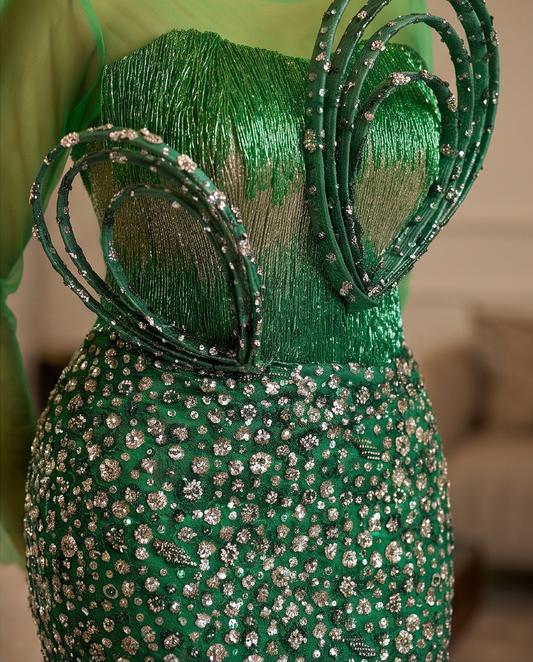 A close-up of a green embellished garment with layered sheer green ribbons and silver rhinestone beadwork, shown against a softly blurred indoor background.