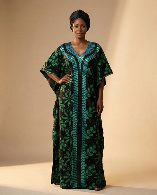 A woman modeling a long black kaftan dress with green floral patterns and teal geometric borders, paired with a black headwrap, against a warm beige studio backdrop.
