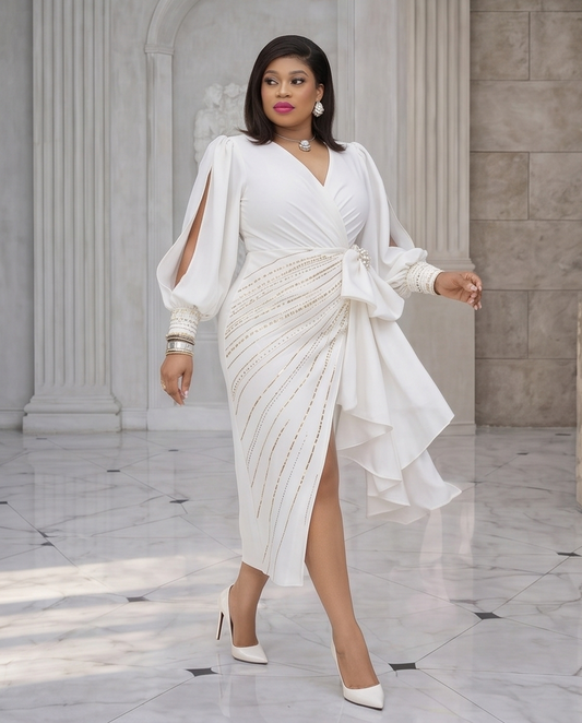 A woman models a white wrap-style midi dress with dramatic draped sleeves, side cutouts, and a sheer ruffled overlay, styled with white pointed heels and jewelry in an elegant marble-floored interior with columns.