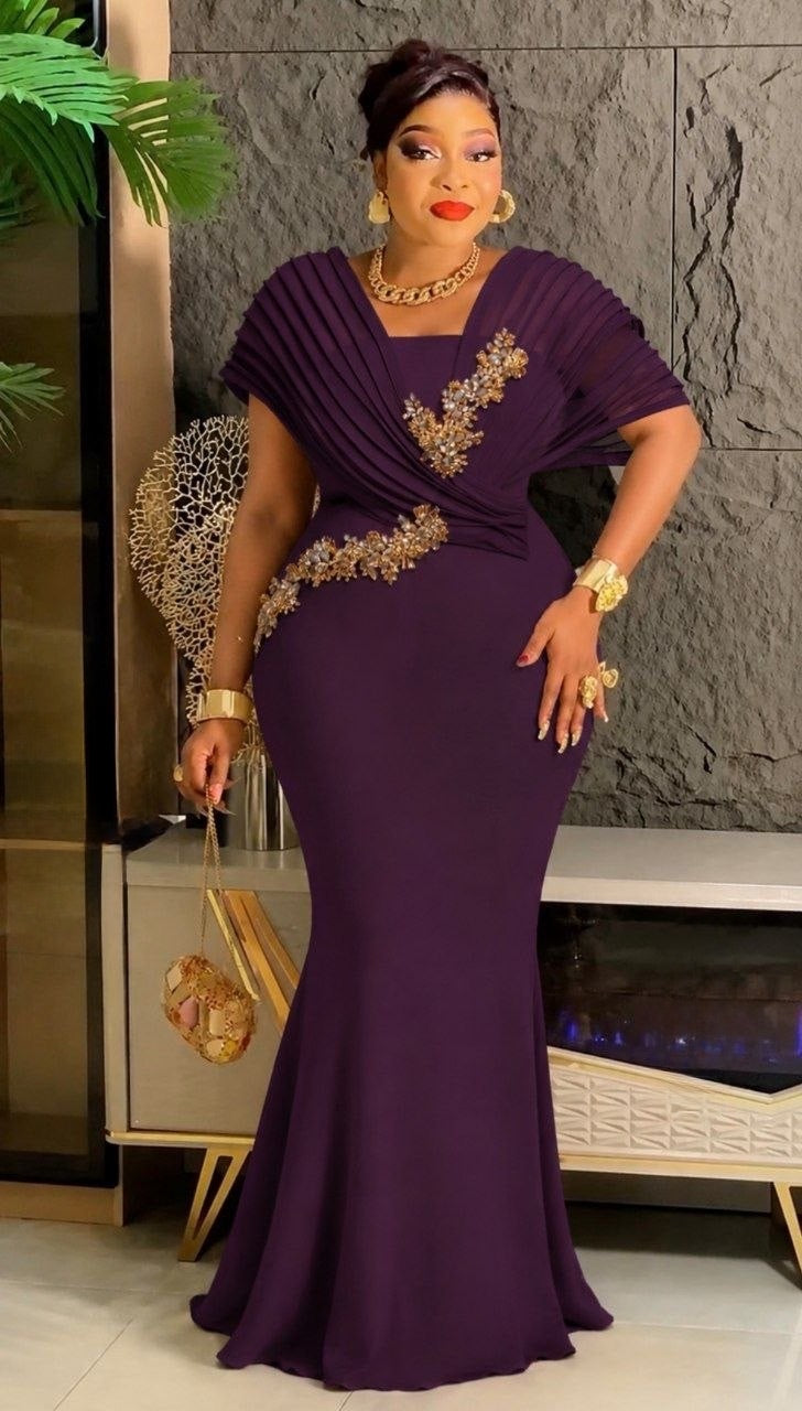 A woman models a floor-length, deep purple mermaid-style gown with draped short sleeves and ornate gold embroidery along the bodice and waist, posed in an indoor setting with a textured gray wall and modern furniture.