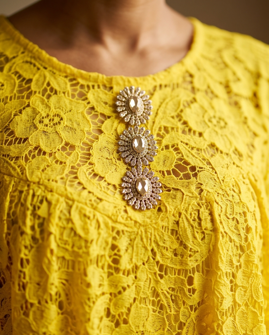 A close-up of a bright yellow lace top featuring three ornate gold-toned brooch embellishments on the chest.