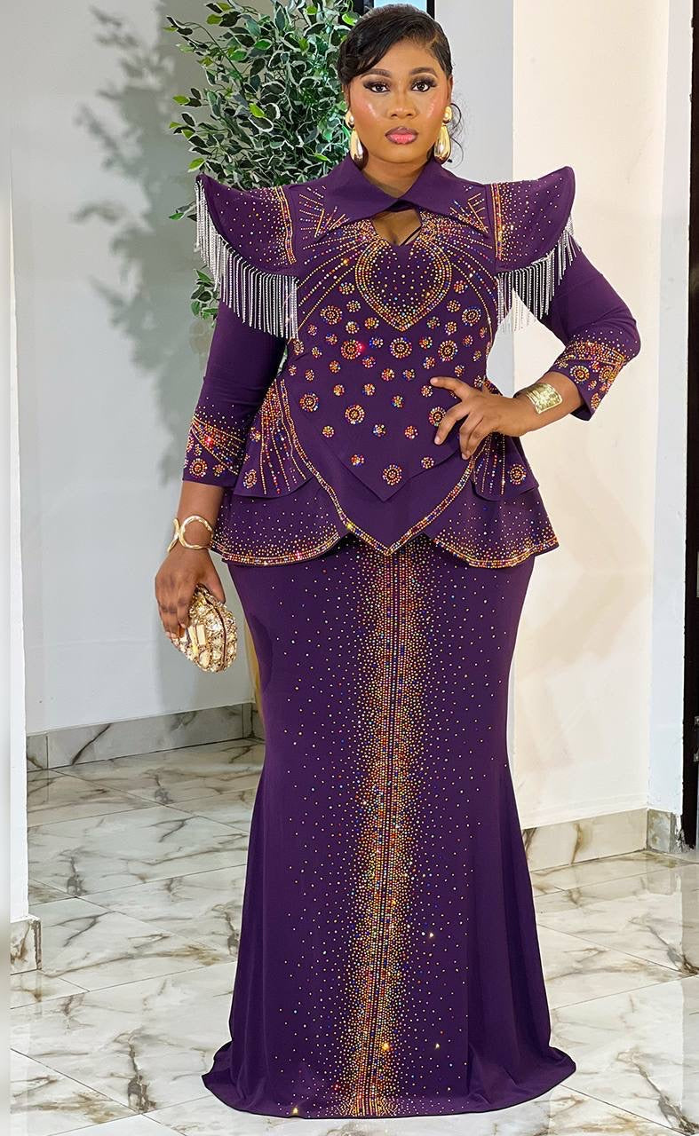 A woman models a purple traditional African dress with gold embroidery, fringe details, and a flared peplum top, standing in an indoor setting with a marble floor.