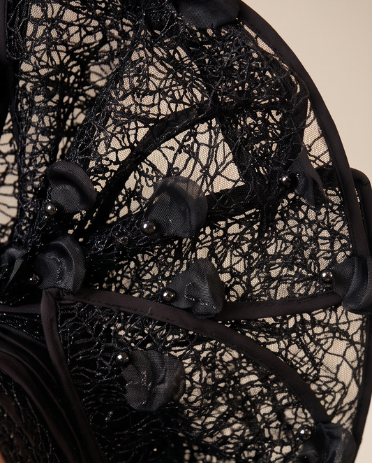 Close-up of a black lace fascinator hat with layered mesh, ribbon loops, and small pearl-like accents against a warm beige background.