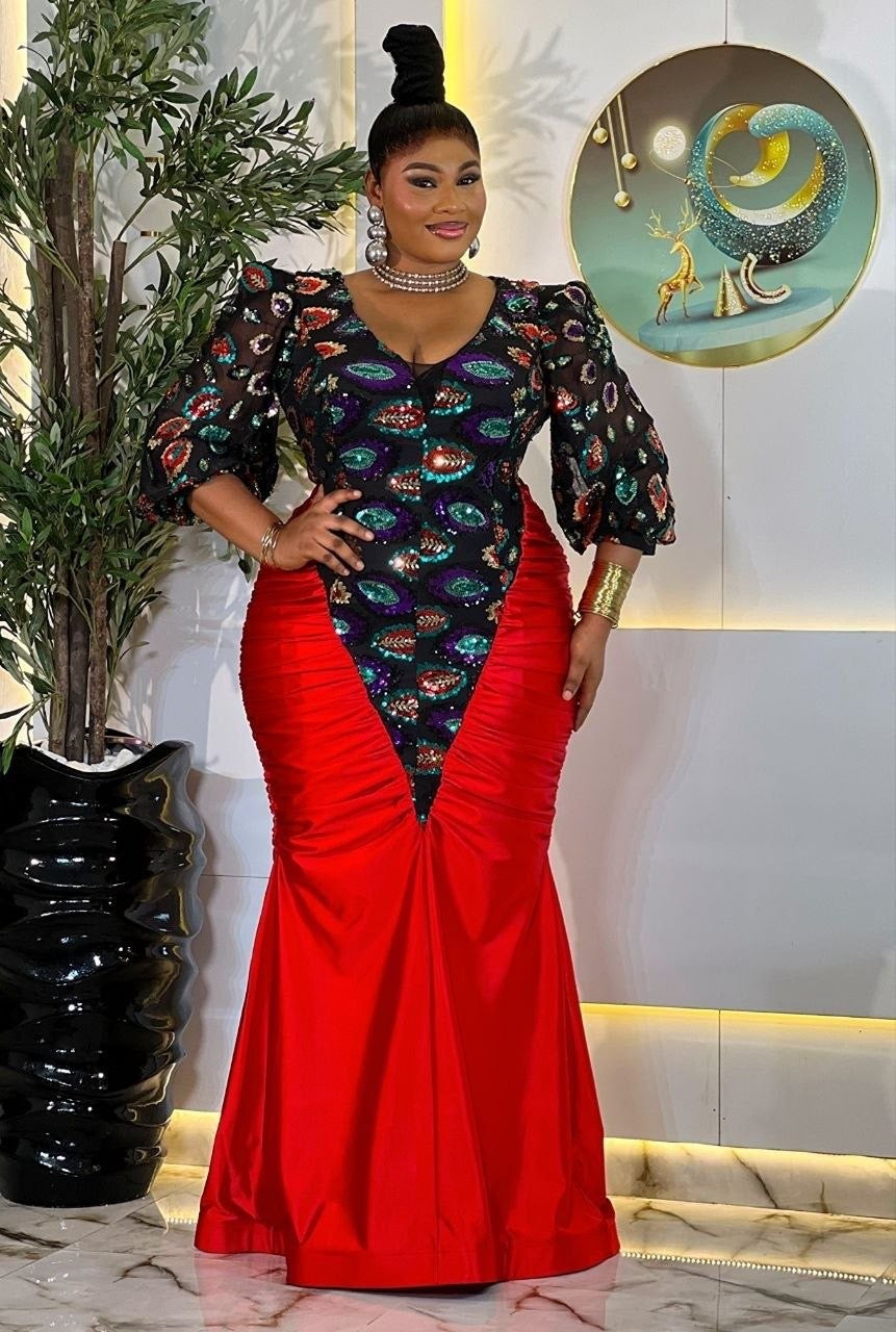 A woman models a floor-length African dress with a vibrant red mermaid skirt and a black bodice decorated with colorful fish prints, set against an indoor background with a plant and wall art.