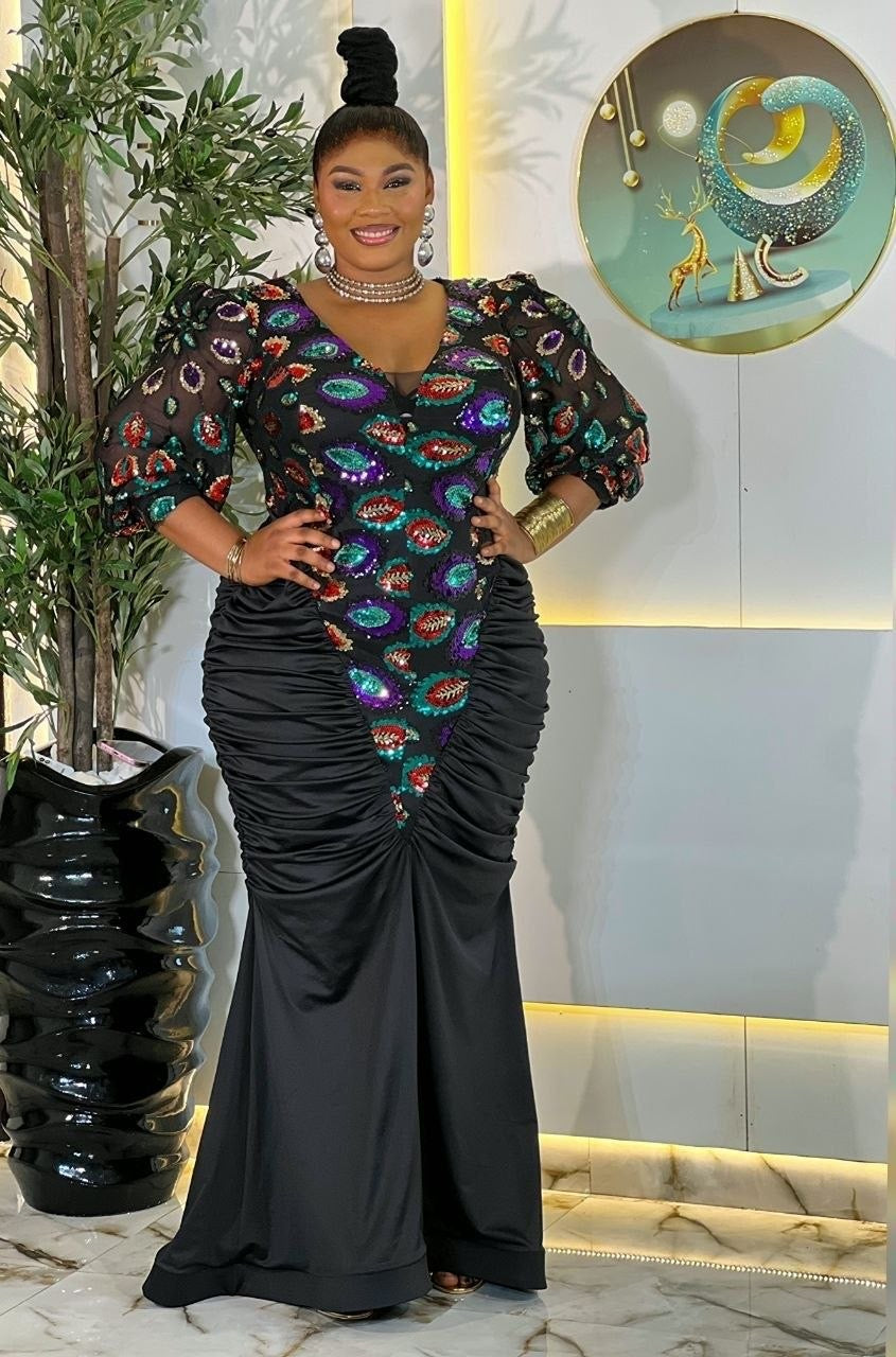 A woman modeling a floor-length black mermaid gown with a heavily embellished multicolor bodice featuring red, green, blue, and purple fish-scale patterns, posed in an indoor setting with a plant and decorative wall art.