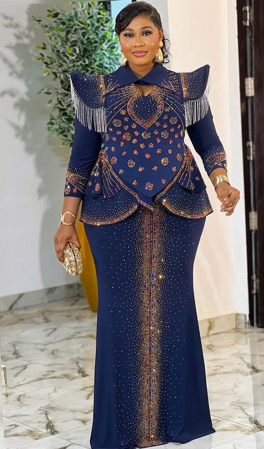 A woman modeling a navy blue two-piece traditional-style dress with gold and orange embroidered details, fringed shoulder accents, and a matching long skirt, standing indoors on a marble floor.