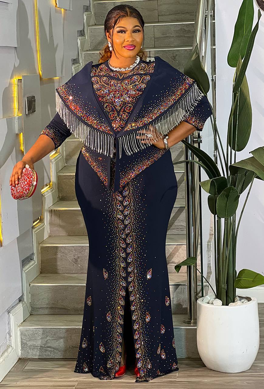 A woman models a dark navy blue floor-length dress with a matching fringed shawl and intricate multicolor embroidered patterns, standing in a modern indoor setting with a staircase and a potted plant.