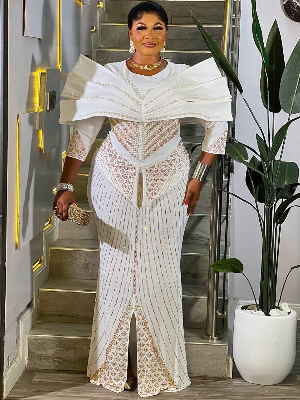Woman in a white and gold dress standing in an indoor setting with a plant and staircase.