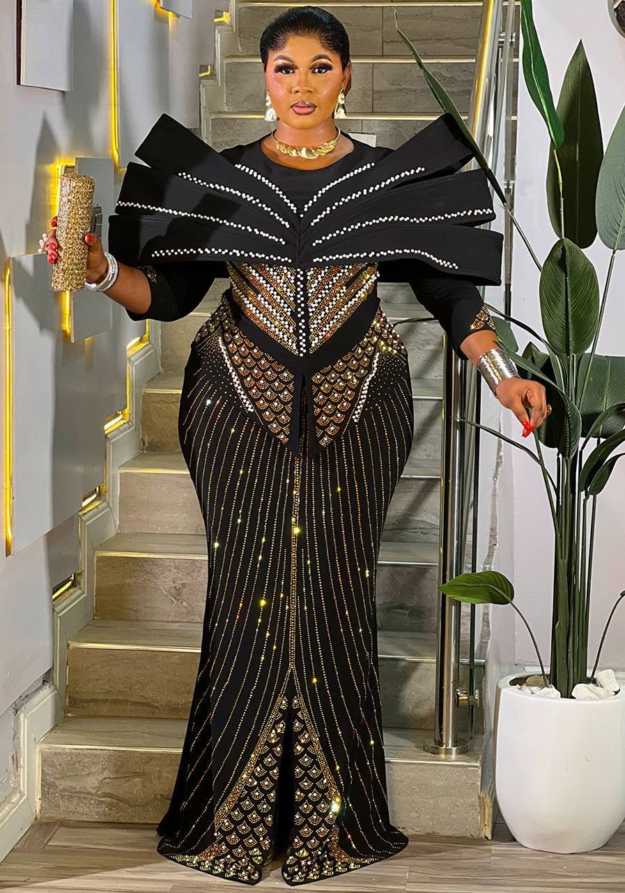 A woman models a black floor-length gown with dramatic layered shoulder ruffles, gold and silver beaded geometric detailing, and vertical gold glitter stripes, standing in a modern indoor staircase setting.
