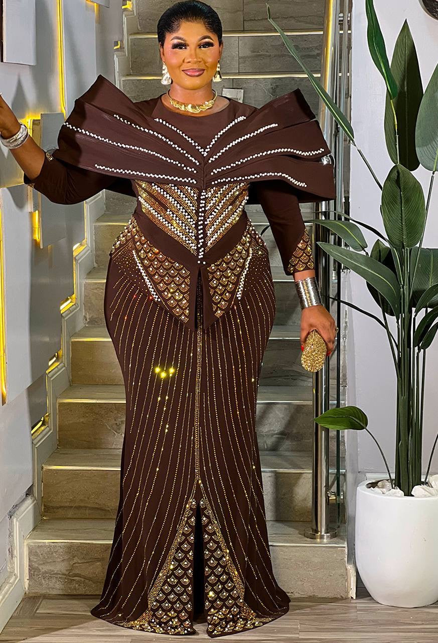 A woman models a floor-length brown gown with dramatic shoulder panels, vertical beaded stripes, and ornate gold embellishments, standing indoors in front of a staircase with a potted plant nearby.