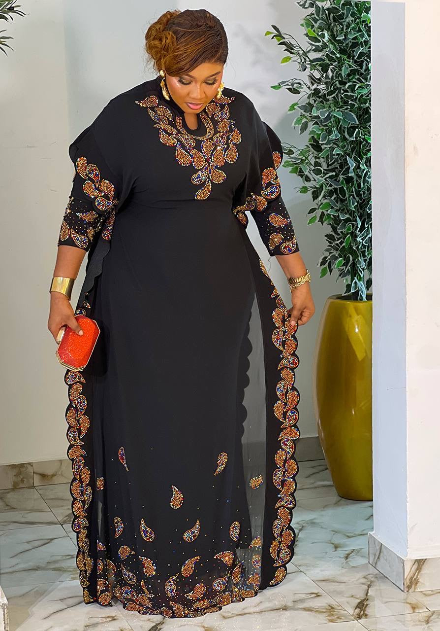 A woman wearing a black maxi dress with orange and brown paisley and floral embroidery, holding a small orange clutch in an indoor setting with a potted plant.