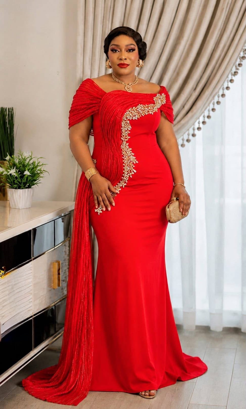 A woman modeling a bright red off-the-shoulder evening gown with gold embroidered detailing along the side, standing in an indoor setting with beige curtains and a white console table.