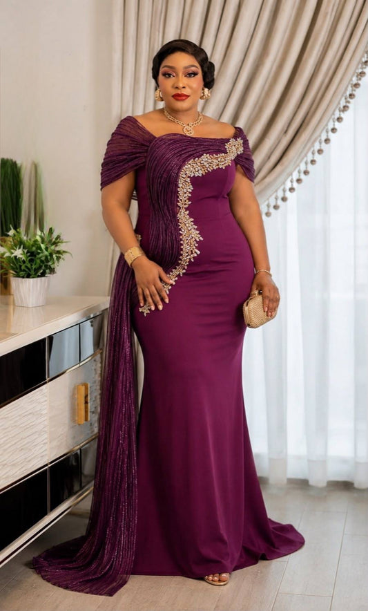 A woman models a floor-length off-the-shoulder gown in deep plum purple with gold sequin embellishments along the side, standing in an indoor setting with beige curtains and a sideboard.