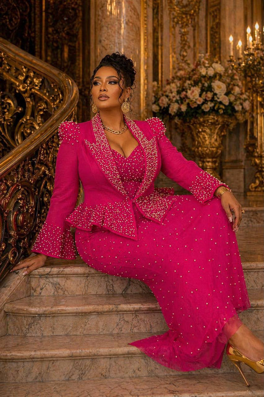 A woman sits on ornate marble stairs in a grand interior, wearing a bright fuchsia embellished gown with a matching jacket and gold heels.